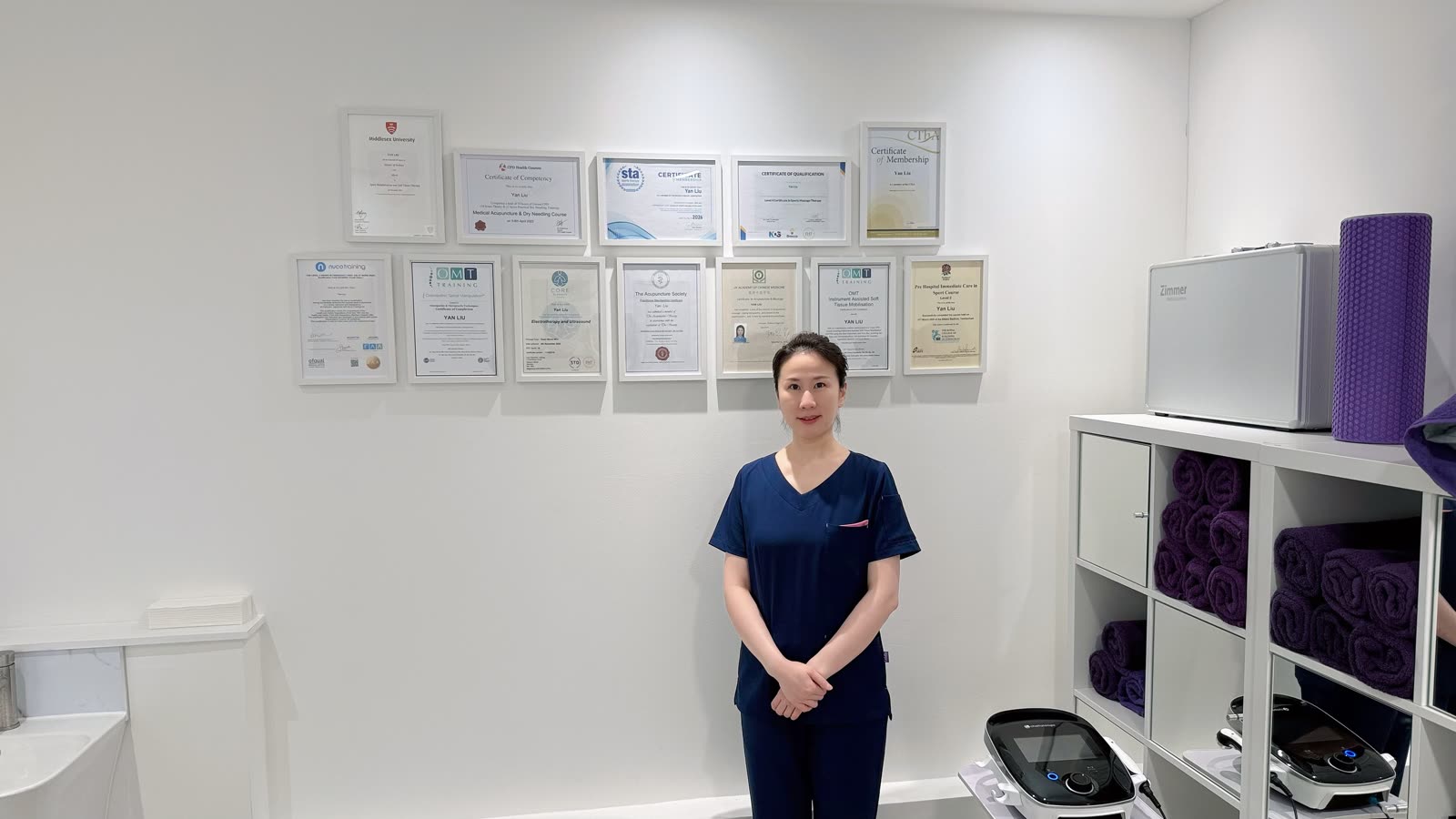 Yan Liu (Joy) in the treatment room at Rehab Joy, standing in front of her framed qualification certificates